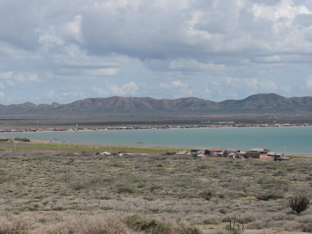 Desde lejos se ve el pequeño pueblo de Cabo de la Vela. En el primer plano se ve un terreno vacío de vegetación y a después el Caribe
